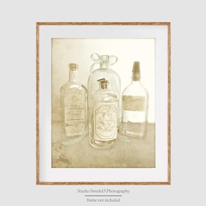 May include: A vintage still life photograph featuring four clear glass bottles with labels and stoppers, arranged on a textured beige surface. The bottles are tied together with a white ribbon.