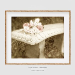 May include: A vintage-style photograph of a white concrete bench with a bouquet of pink and white roses on top. The background is blurred and has a sepia tone.