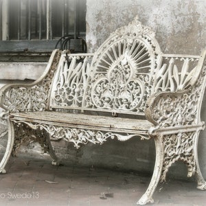May include: A white cast iron bench with intricate floral designs. The bench is weathered and has a rustic look. The bench is sitting in front of a white wall.