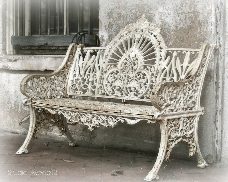 May include: A white cast iron bench with intricate floral designs. The bench is weathered and has a rustic look. The bench is sitting in front of a white wall.