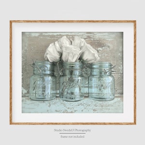 May include: Three vintage glass jars with white flowers in the center jar, on a light blue and brown wood background.