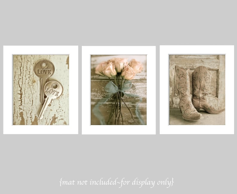 Op de afbeelding: Set van drie ingelijste prints met een rustiek thema. De eerste print toont twee sleutels met het woord "LOVE" erop. De tweede print toont een boeket roze rozen gebonden met een blauw lint. De derde print toont een paar bruine cowboylaarzen.