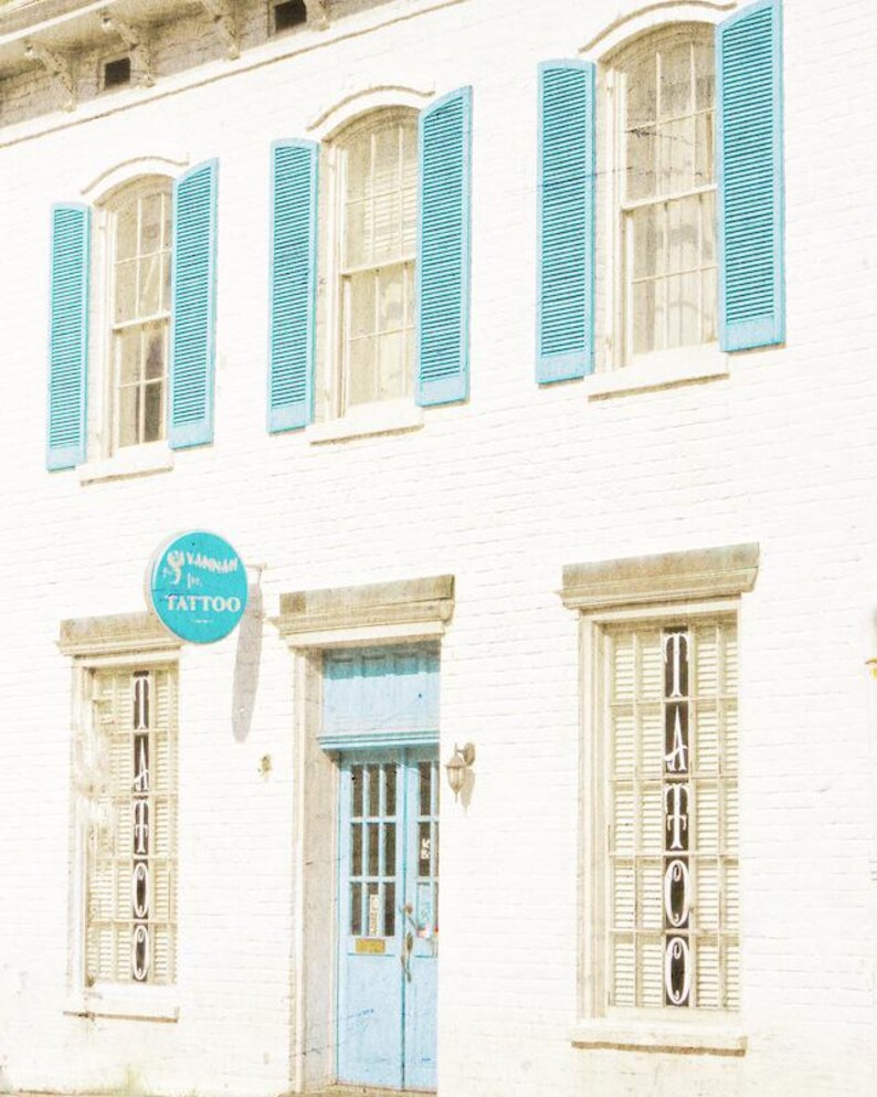 May include: A white building with blue shutters and a blue door. The building has a sign that says "Glamour Ink Tattoo".
