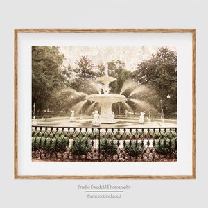 May include: A vintage-style photograph of a white fountain with water spraying out in a park setting. The fountain is surrounded by a black wrought iron fence and lush green foliage. The image has a faded, sepia tone.