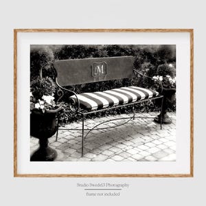 May include: Black and white photo of a metal bench with a striped cushion. The bench has an initial "M" on the back. The bench is in a garden setting with potted plants.