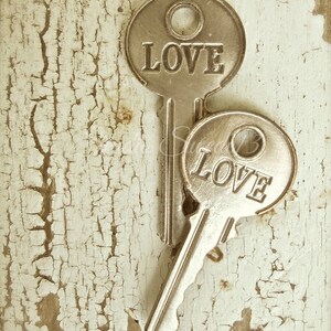 Op de afbeelding: Twee antieke zilveren sleutels met het woord "LOVE" gegraveerd op elk. De sleutels liggen op een wit, gebarsten houten oppervlak.