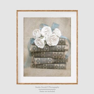 Puede incluir: Una pila de libros antiguos con un ramo de rosas blancas y una cinta azul en la parte superior. Los libros tienen una cubierta de cuero marr&oacute;n con detalles dorados. El fondo es un beige suave y descolorido.