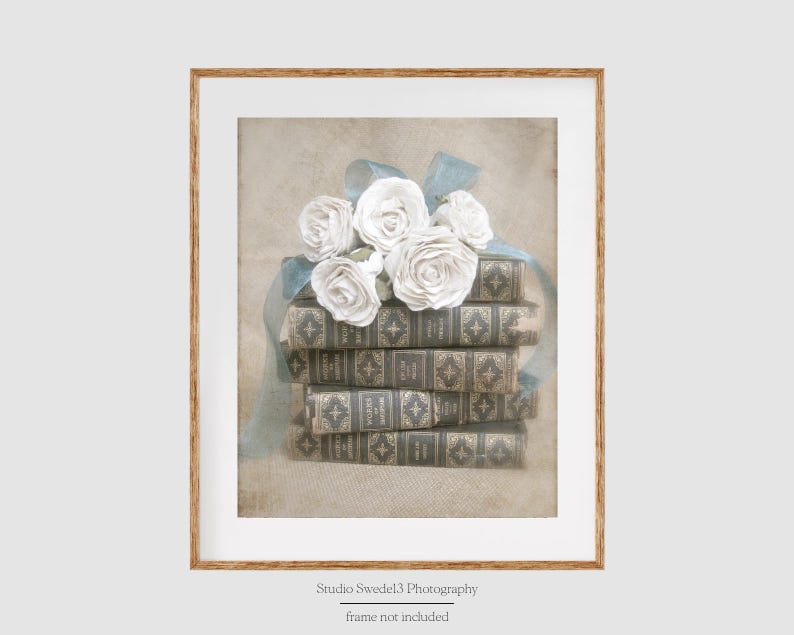 Puede incluir: Una pila de libros antiguos con un ramo de rosas blancas y una cinta azul en la parte superior. Los libros tienen una cubierta de cuero marr&oacute;n con detalles dorados. El fondo es un beige suave y descolorido.