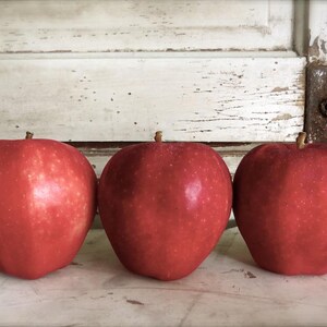May include: Three red apples on a white wooden surface. The apples are arranged in a row, with the largest apple in the middle.