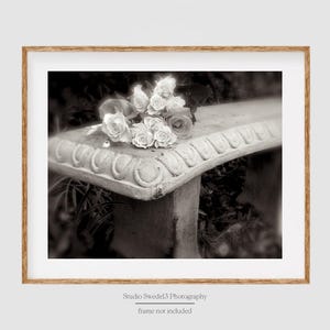 May include: Black and white photograph of a stone bench with a bouquet of roses. The bench has ornate detailing along the top edge. The roses are clustered together, with some blooms fully open. The photograph is framed in a wooden frame.