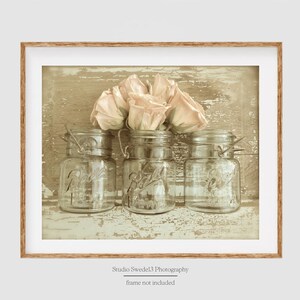 May include: Three clear glass jars with metal lids filled with pale pink roses on a distressed wood background.