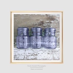 May include: Three vintage glass jars with purple tint and wire lids, sitting on a weathered wooden surface with white paint.