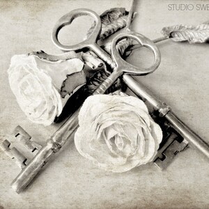 May include: Two antique keys with ornate designs are intertwined with two white fabric roses. The keys are silver and have a vintage look. The roses are delicate and soft, adding a romantic touch to the image.