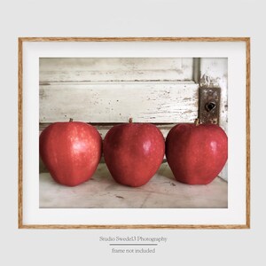 May include: Three red apples lined up in a row on a white, weathered wooden surface. The apples are in focus, while the background is slightly blurred.