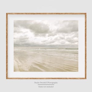 May include: A framed photograph of a beach scene with a cloudy sky and waves crashing on the shore. The sand is light brown and the water is a light blue.