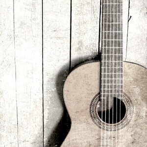 Op de afbeelding: Een close-up van een houten akoestische gitaar met een lichtbruine afwerking. De gitaar leunt tegen een witte houten muur. De gitaar heeft een zwart klankgat met een decoratieve inlay.