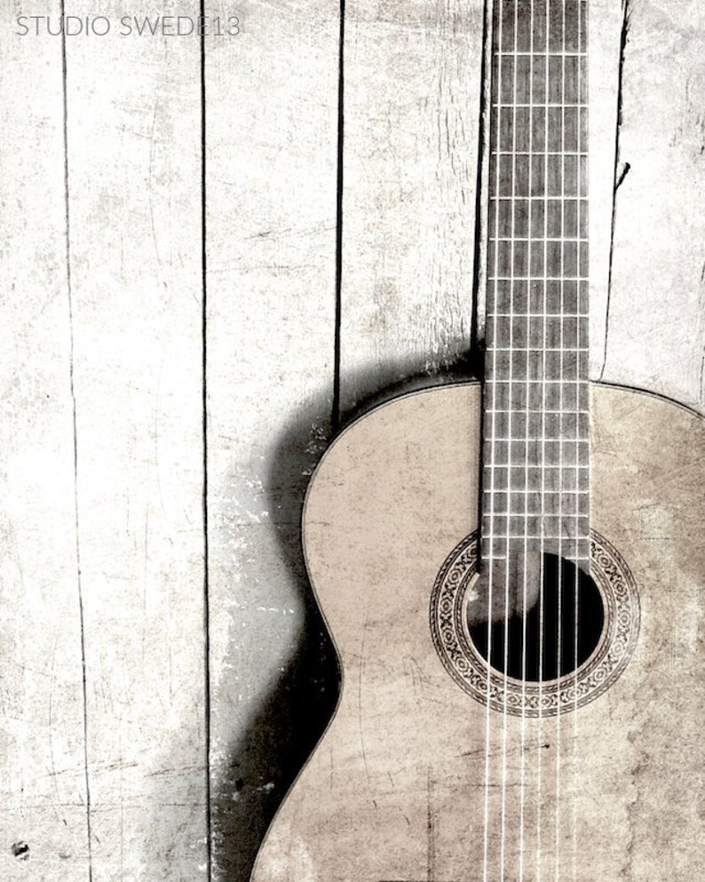 Op de afbeelding: Een close-up van een houten akoestische gitaar met een lichtbruine afwerking. De gitaar leunt tegen een witte houten muur. De gitaar heeft een zwart klankgat met een decoratieve inlay.
