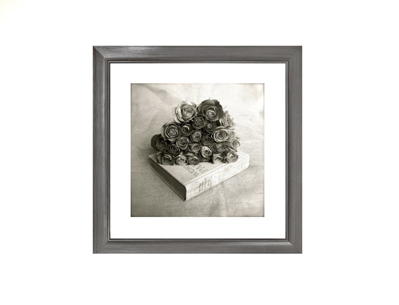 May include: A black and white photograph of a bouquet of roses on top of a closed book. The book is titled "The World of Interiors". The image is framed in a gray frame.