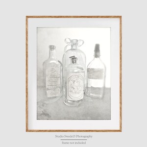 May include: A black and white photograph of four glass bottles with labels. The bottles are arranged in a row on a white background. The bottles are all different shapes and sizes. The photograph is framed in a light wood frame.