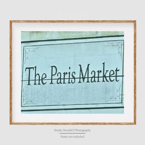 May include: A blue and white sign with ornate detailing reads "The Paris Market" in black lettering.
