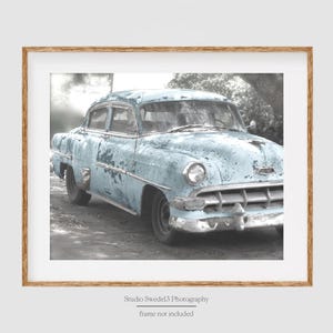 May include: A vintage light blue car with faded paint and chrome accents. The car is parked on a gray asphalt surface. The background is blurred and out of focus.