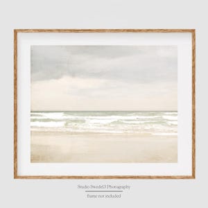 May include: A framed photograph of a beach scene with white sand, blue water, and a cloudy sky. The image is in a light wood frame.