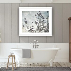 May include: A framed photograph of a field of tall, dried wildflowers against a cloudy sky. The photo is in a white frame and is hanging on a white wall in a bathroom.