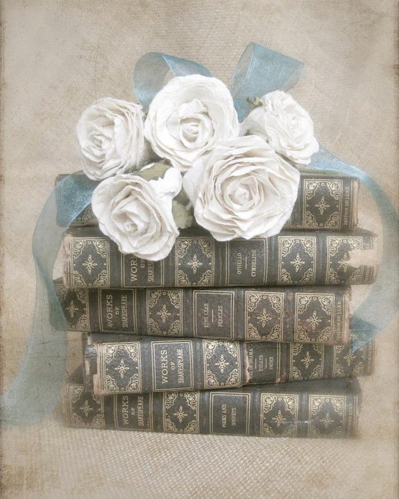 Puede incluir: Una pila de libros antiguos con letras doradas y dise&ntilde;os ornamentados en las cubiertas. Un ramo de rosas de papel blanco con una cinta azul se coloca encima de los libros.