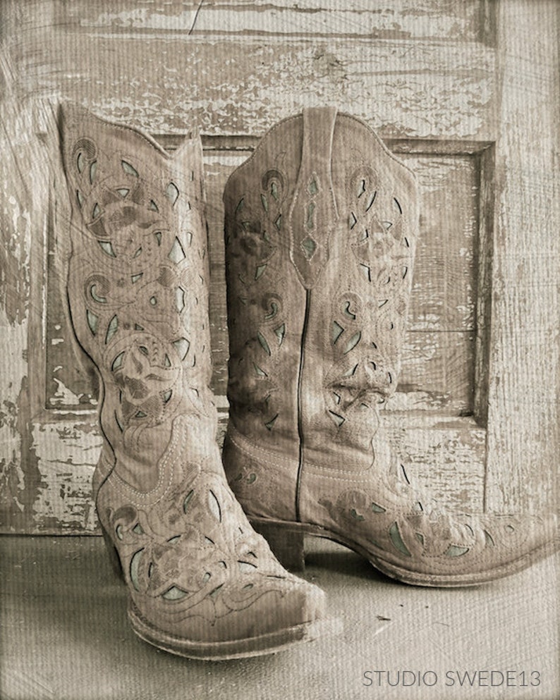 Op de afbeelding: Een paar lichtbruine cowboylaarzen met ingewikkelde uitgesneden ontwerpen. De laarzen staan tegen een rustieke houten achtergrond met een verweerde witte verfbehandeling.