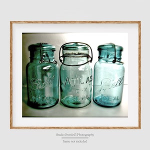 May include: Three vintage blue-green glass jars with metal closures. The jars are labeled "Ball Ideal" and "Atlas E-Z Seal". The jars are displayed in a wooden frame. The image is a photograph.