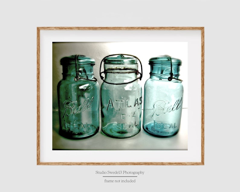 May include: Three vintage blue-green glass jars with metal closures. The jars are labeled "Ball Ideal" and "Atlas E-Z Seal". The jars are displayed in a wooden frame. The image is a photograph.