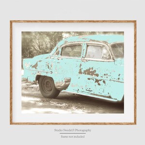 May include: A vintage, light blue car with rust and faded paint. The car is parked on a dirt road with trees in the background.