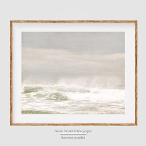 May include: A framed photograph of a seascape with white foamy waves crashing against the shore. The sky is a soft, light blue with wispy clouds. The image has a vintage, faded look.