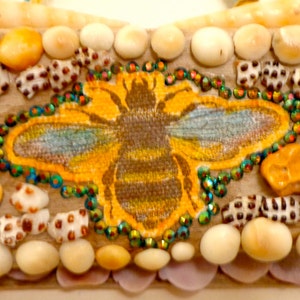 May include: A close-up of a bee mosaic made with seashells and rhinestones. The bee is yellow and brown with blue wings. The mosaic is surrounded by a border of white and brown seashells.