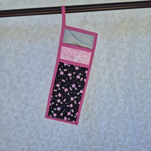 May include: A quilted oven mitt with a pink flamingo pattern on a black background. The mitt has a pink border and a hanging loop. The design includes sections of pink and gray fabric. The mitt is hanging from a brown rod.