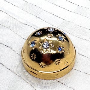 May include: A gold-colored, dome-shaped trinket box adorned with star and floral designs. The stars are embellished with blue and clear gemstones. The box is set against a white, textured background.