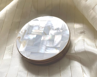 May include: A round, decorative box with a mosaic-style lid made of iridescent white and cream-colored pieces. The box has a bronze-colored metal rim and sits on a cream-colored fabric with gold stripes.