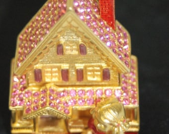 May include: A gold-colored house figurine with pink rhinestones and a red chimney. A small pink figurine of a girl is sitting in front of the house.