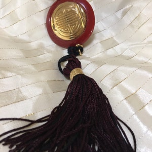 May include: A red and gold pendant necklace with a tassel. The pendant is round and features a gold Chinese symbol. The tassel is dark brown.