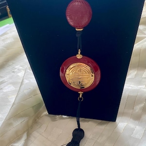 May include: A red and gold pendant necklace with a black cord and tassel. The necklace features two red, round, carved pendants with gold accents. The lower pendant has a gold emblem. The necklace is displayed on a black stand.