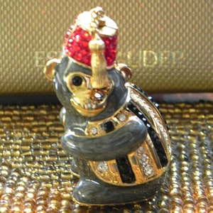 Puede incluir: Una pequeña figurita de esmalte gris de un mono que lleva un gorro rojo y dorado con borla. El mono lleva un chaleco a rayas blancas y negras con ribetes dorados y diamantes de imitación. La figurita está sentada sobre un fondo dorado.