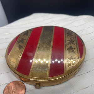 May include: A round, vintage compact with a red and gold striped design. The compact has a gold-colored metal frame and is decorated with leaf patterns. A penny is next to the compact for size comparison.