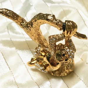 Puede incluir: Una figura decorativa dorada de una ardilla colgando de una rama. La ardilla está adornada con cristales transparentes, añadiendo brillo. La rama tiene una apariencia texturizada similar a la corteza, y la figura tiene ojos negros.