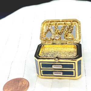 May include: A small, open jewelry box with a gold exterior and navy blue accents. The box has two small drawers and a hinged lid. Inside the lid are small gold chains with decorative charms. A penny is next to the box.