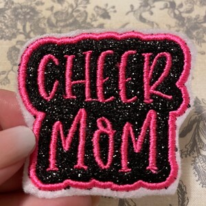 May include: Black and pink glitter iron-on patch with the text "Cheer Mom" in a white outline.