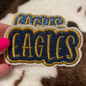 May include: A blue and gold glitter iron-on patch with the word "Eagles" in a cursive font.