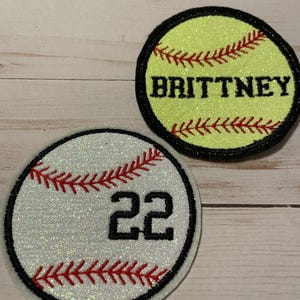 May include: Two white and yellow softball patches with red stitching. One patch has the name "BRITTNEY" embroidered on it. The other patch has the number "22" embroidered on it.