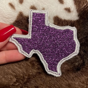 May include: Purple glitter iron-on patch shaped like the state of Texas with a white outline.