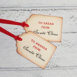 Puede incluir: Dos etiquetas de regalo de estilo vintage con cinta roja. Las etiquetas son de color crema con bordes marrones y tienen el texto "TO: SARAH FROM: Santa Claus" y "TO: AMANDA FROM: Santa Claus" escrito en cursiva negra.
