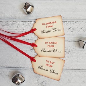 Puede incluir: Tres etiquetas de regalo de estilo vintage con cinta roja. Las etiquetas son marrones con un aspecto desgastado y tienen el texto "TO: AMANDA FROM: Santa Claus", "TO: SARAH FROM: Santa Claus" y "TO: ROY FROM: Santa Claus" impreso en rojo.